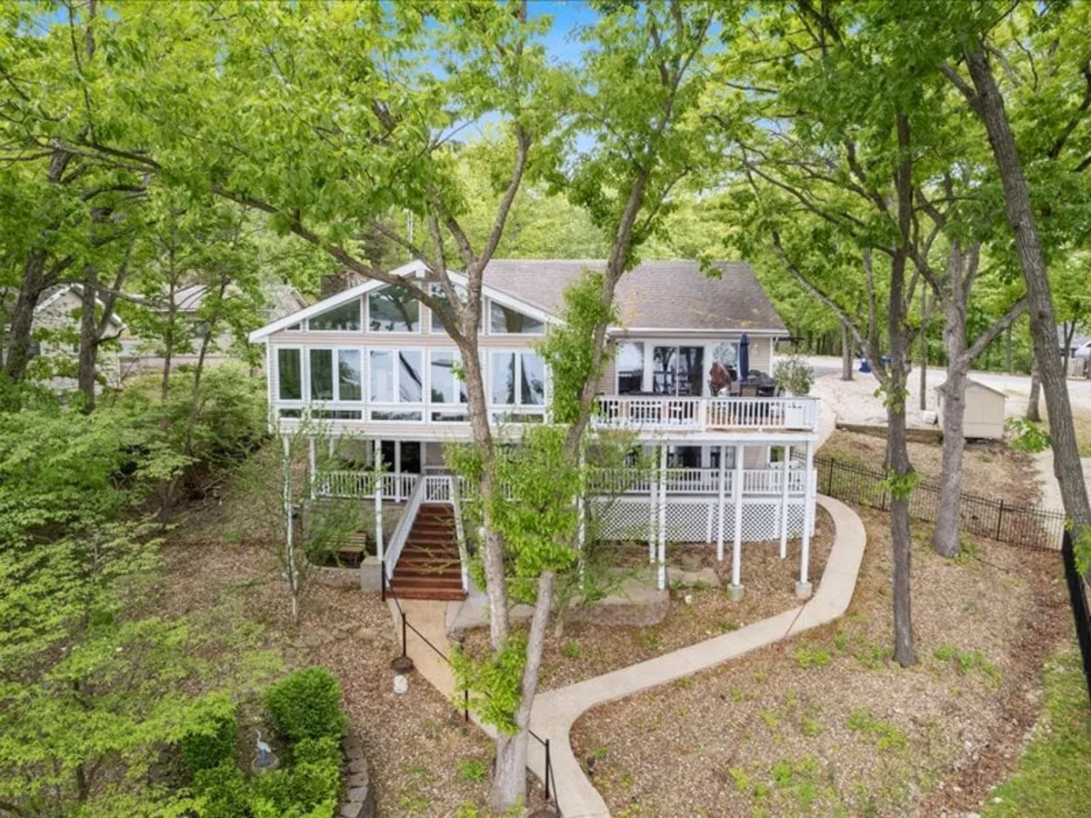 The image features large glass windows opening onto a wraparound porch surrounded by a landscaped garden. The porch is designed for relaxation and entertaining, offering panoramic views of the natural surroundings. The scene captures the relaxing, resort-like ambiance of the property.