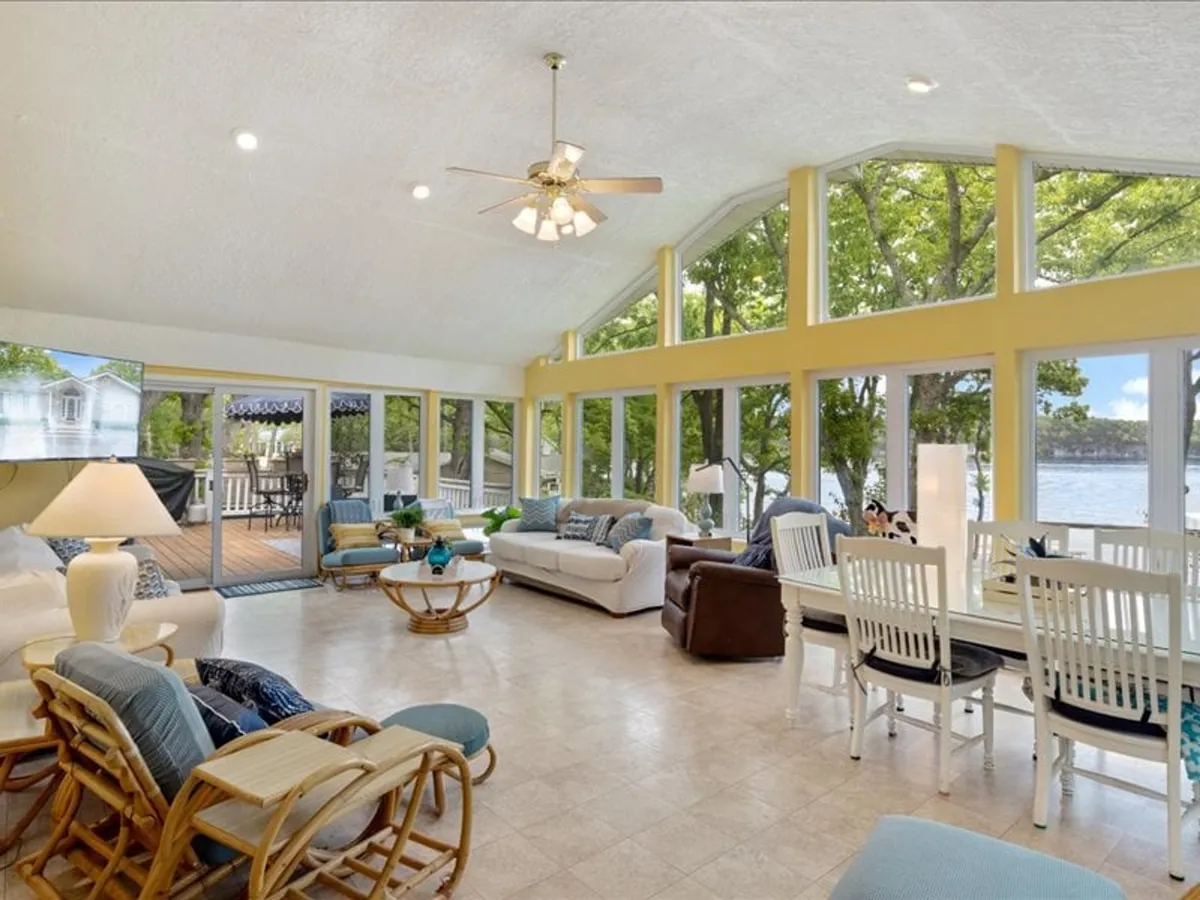 A spacious living area with large windows overlooking the water, featuring a comfortable seating area with plush sofas and a dining table with chairs. Soft natural light streams in, illuminating the space and highlighting the serene views of the lake beyond. Decorative touches like a lamp and cozy furnishings reinforce the inviting atmosphere.