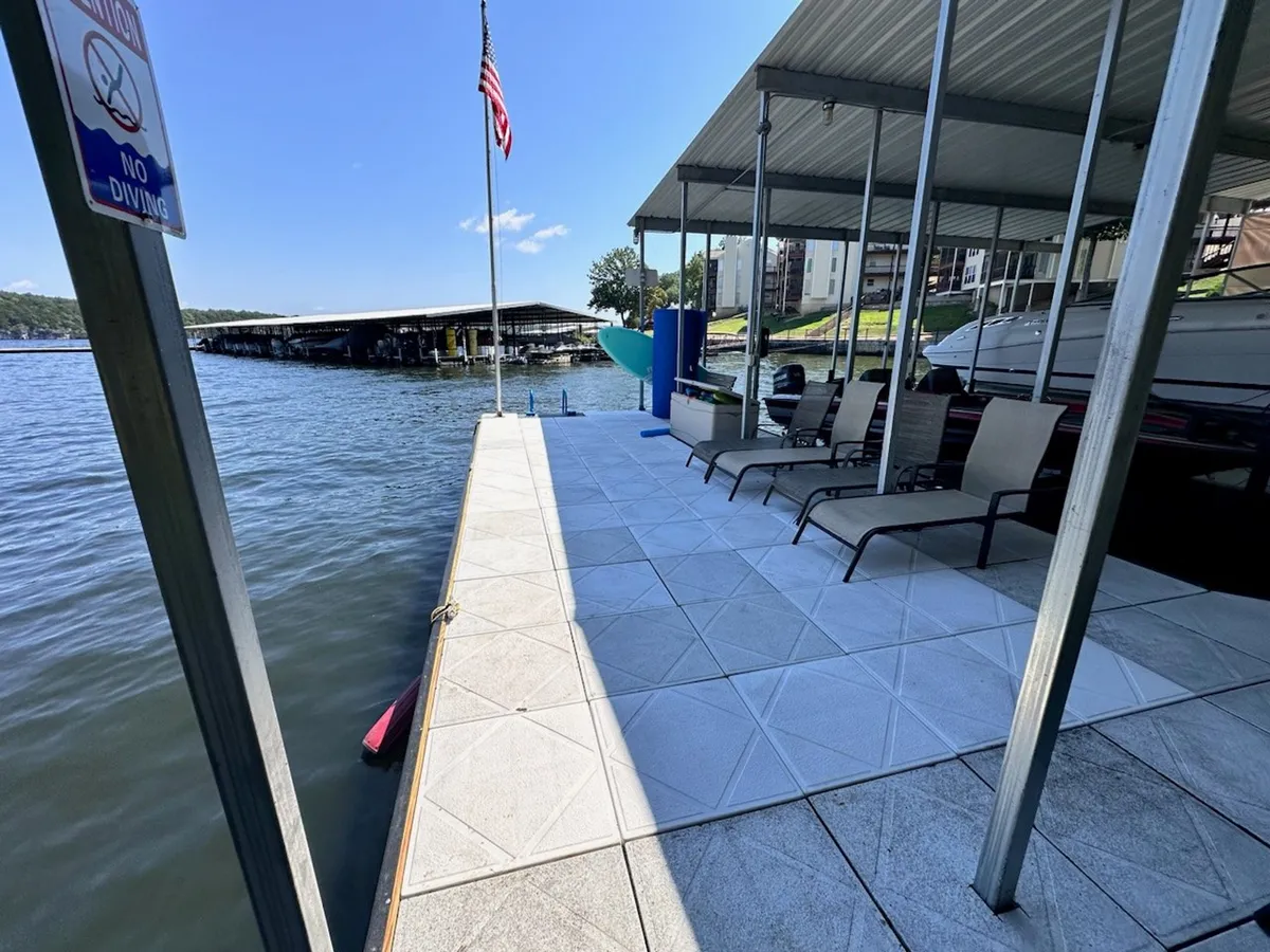 A docking area with lounge chairs facing the water and an American flag gently waving. The water surface glimmers in the sun, and the dock is clearly designed for both fishing and relaxing, with space to gather or enjoy a quiet moment.