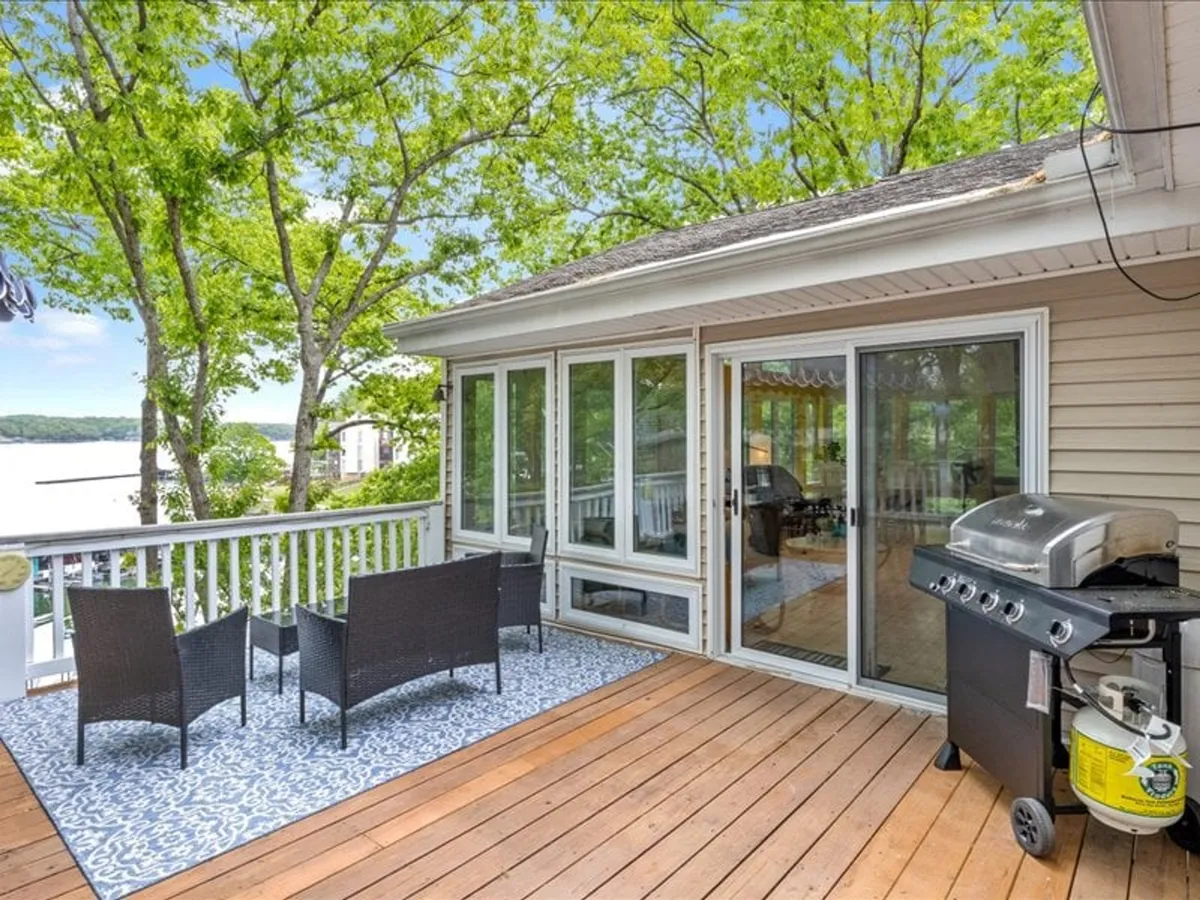 A stylish outdoor seating area with a grill overlooks the serene lake. The spacious deck is arranged for gatherings, with comfortable seating and a clear view of the water, perfect for sunset dinners, yoga, or stargazing. Lush greenery and the tranquil lake make it ideal for both relaxation and socializing.
