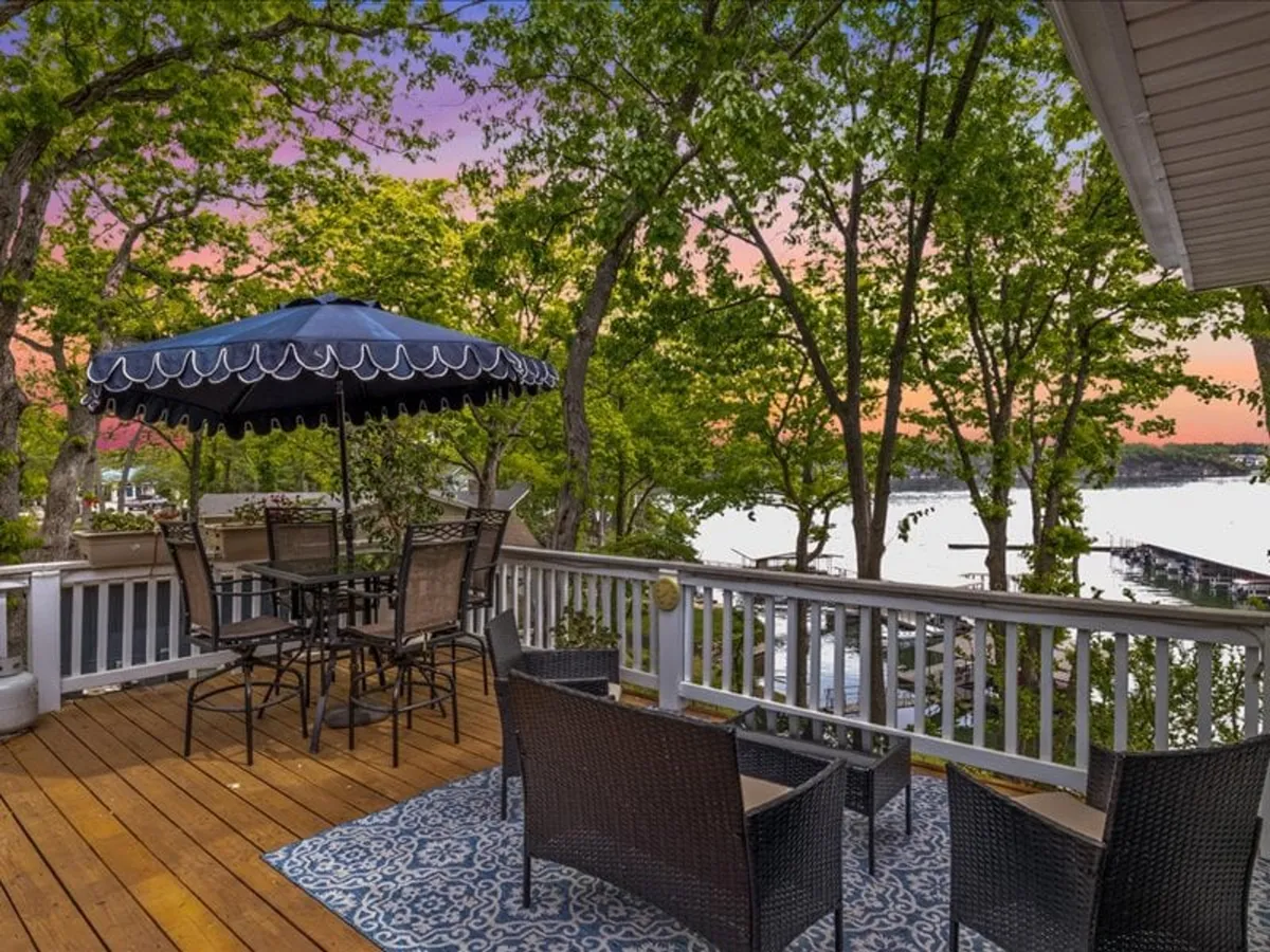 An inviting patio setup with a table and umbrella, woven chairs, a decorative rug, and direct sightlines to the lush greenery and water beyond. The scene is set for outdoor dining or relaxing while enjoying natural beauty and waterfront vistas.