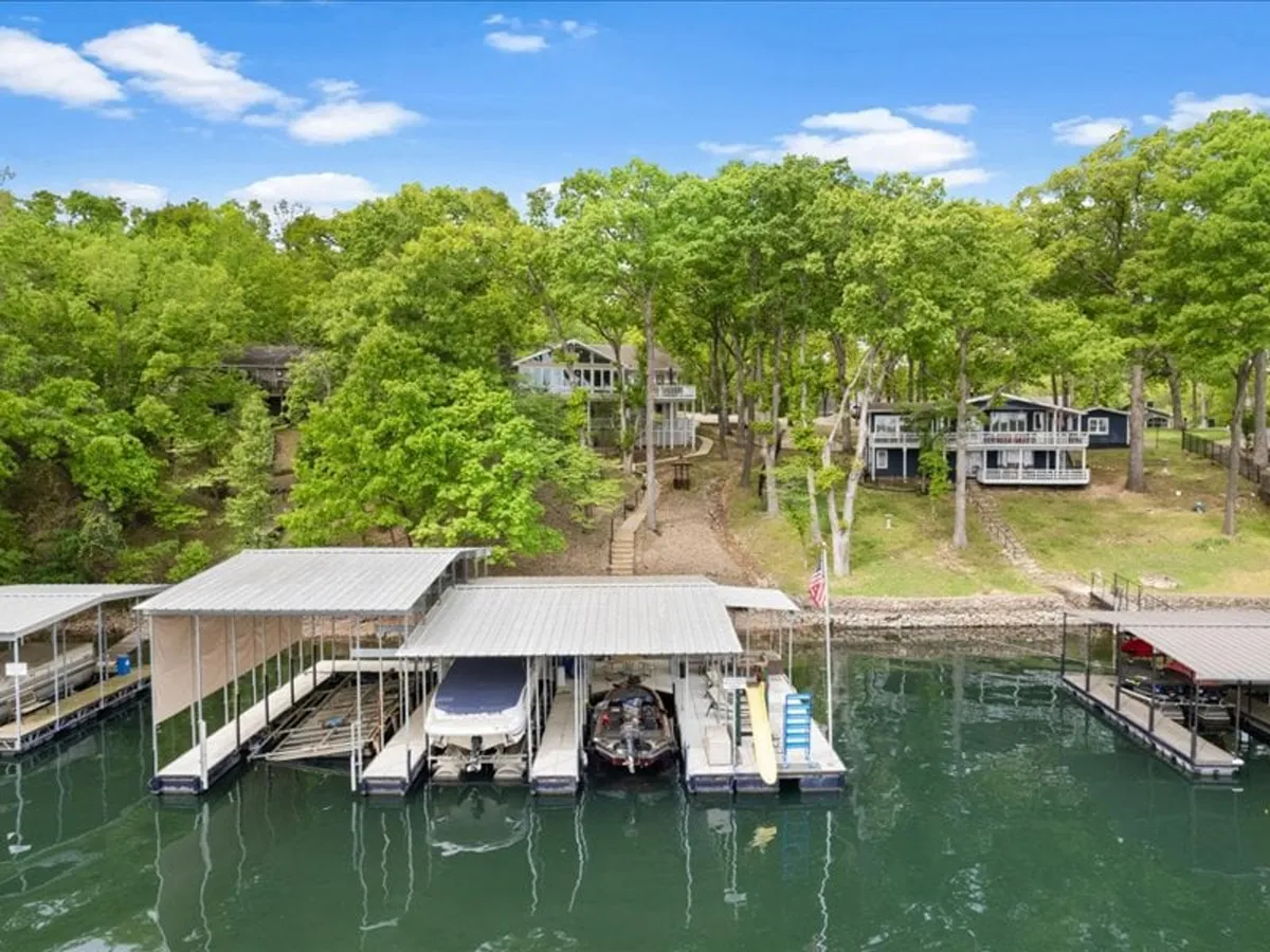 A serene lakeside view featuring a dock with boats moored alongside, lush green trees lining the shore, and residential houses nestled on the hillside. The tranquil water reflects the warm hues of a sunset, capturing the peaceful spirit of Osage Beach.