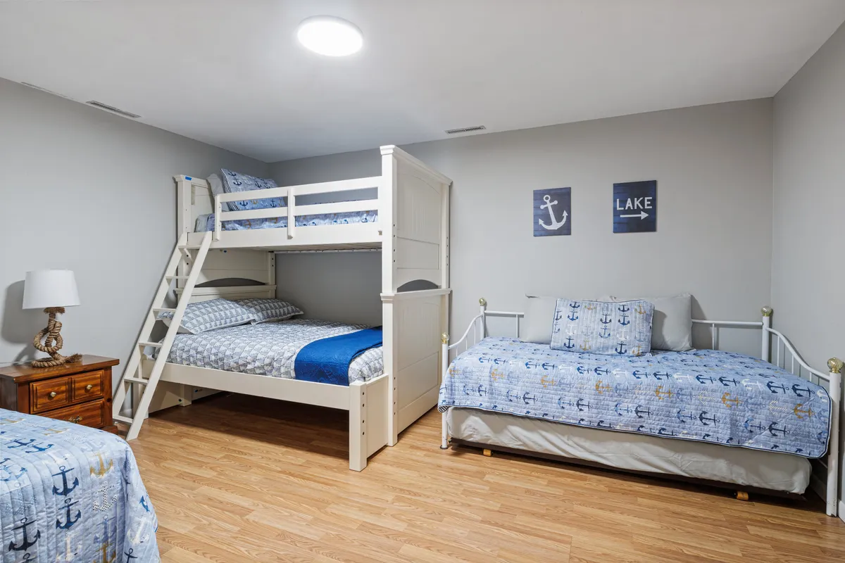 A playful and relaxed bedroom featuring a bunk bed and single bed with nautical-themed bedding, anchor and lake wall art, and a bedside lamp. The layout is perfect for children or friends to enjoy sleepovers and family bonding.