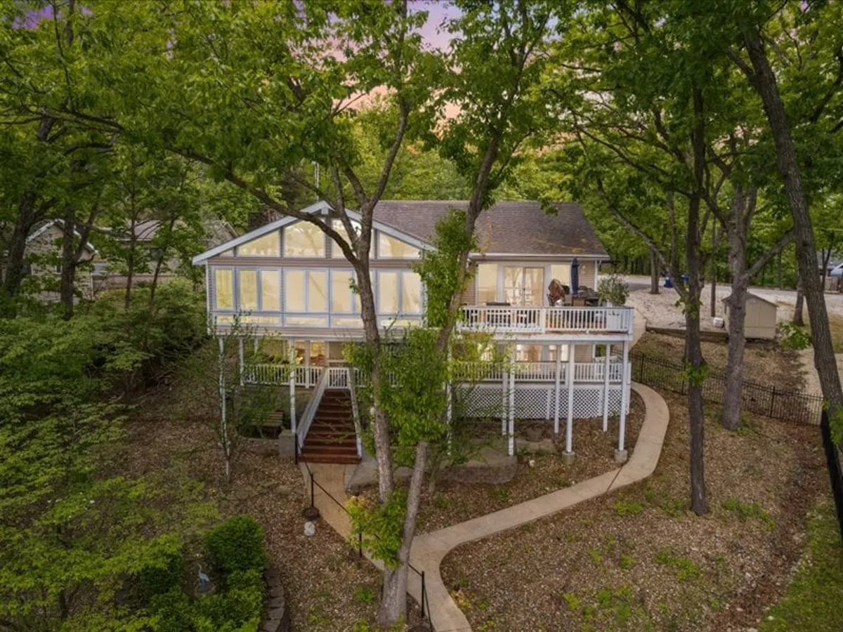 The image showcases the exterior of a spacious lakefront home with a welcoming balcony overlooking serene water views. A well-kept path leads to the entrance, inviting guests to step inside. The balcony is set up for relaxation or gatherings, framed by lush greenery and the tranquil backdrop of the lake.
