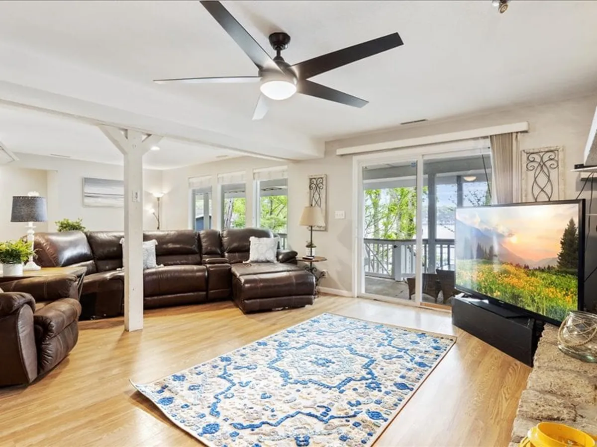 A cozy living room anchored by a large leather sectional sofa, a decorative rug, and a big TV, with sliding doors opening to outdoor views. The setup is perfect for movie nights, family game time, or simply watching the sunset from the comfort of indoors. The balance of soft interiors and visible lake views through the glass connects the indoor experience to the natural surroundings.
