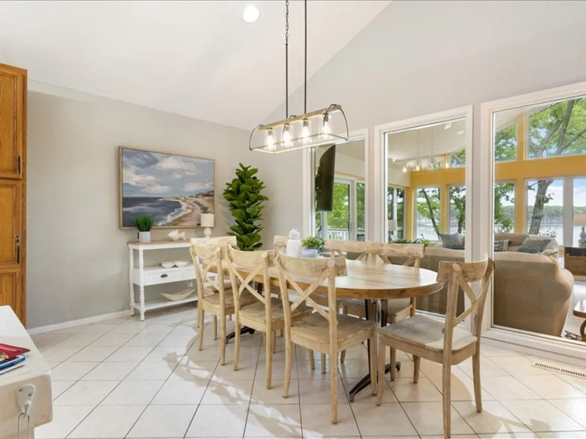 A bright dining area featuring a wooden table, large windows overlooking the water, green plant decor, and wall art. The room feels open, airy, and perfect for group gatherings, with natural light enhancing the inviting atmosphere.