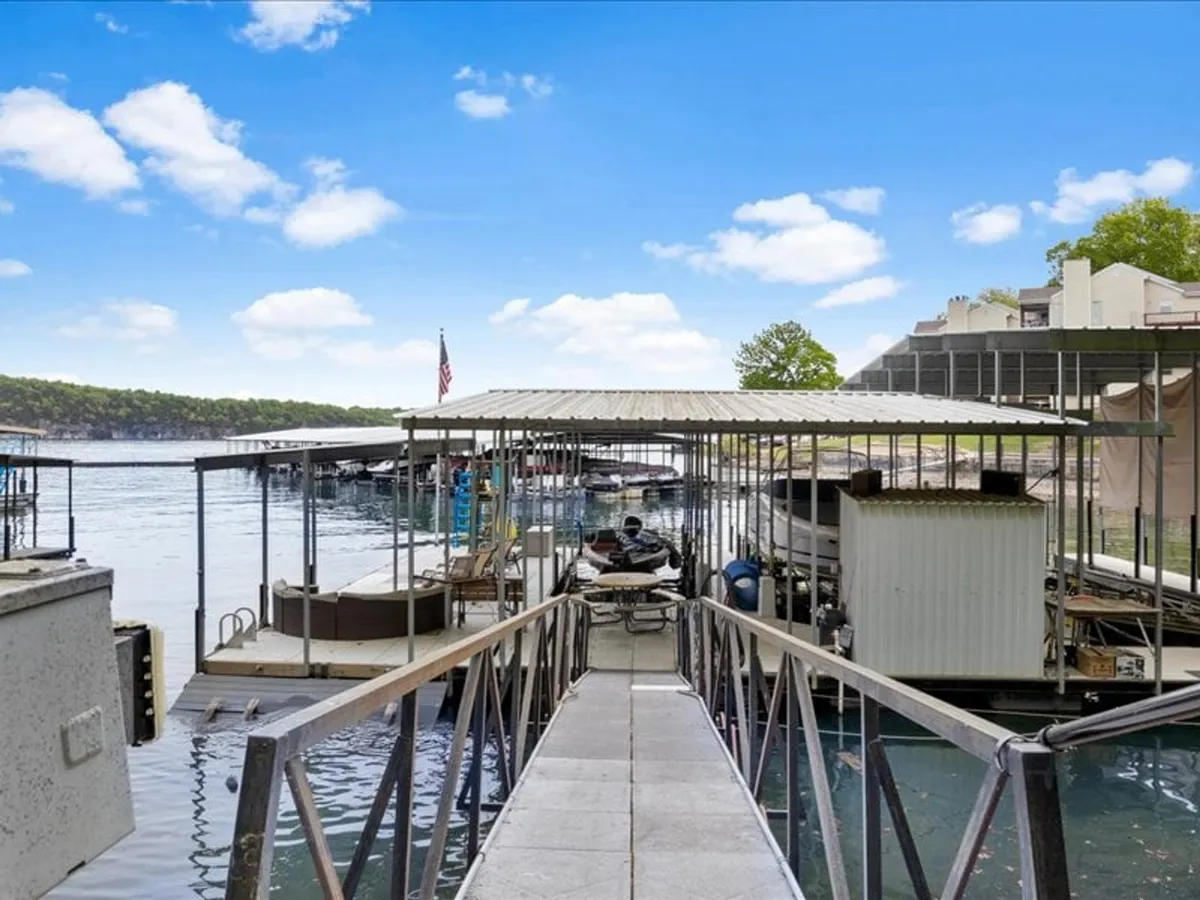 This image features a tranquil lakeside scene with a dock extending over calm water, surrounded by boathouses, trees, and an expansive sky. The dock and water are central, inviting viewers to imagine fishing right from the edge, while the natural setting underscores a peaceful, recreational atmosphere.