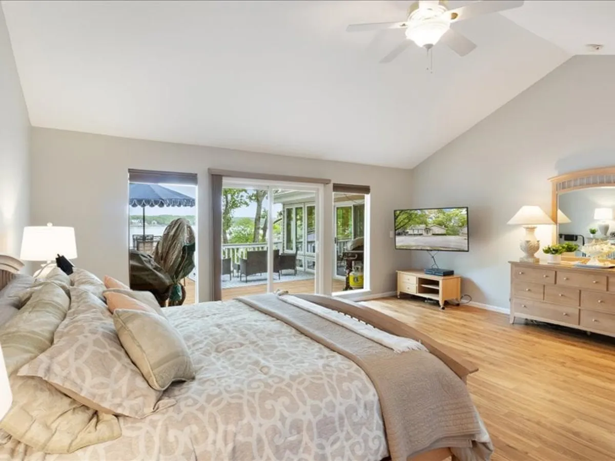 A serene bedroom with a large bed adorned with decorative pillows, expansive windows overlooking the deck and lake, a media console with television, and a wooden dresser decorated with homey touches. Natural light floods the room, emphasizing the tranquil atmosphere and lake views.