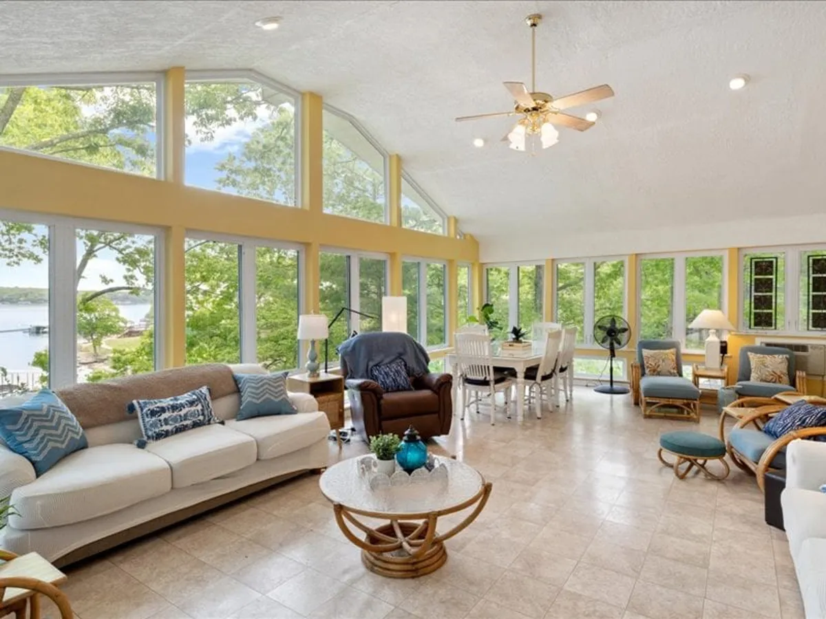 A bright, welcoming open-concept living and dining room with large windows offering panoramic views of the outdoors. Comfortable seating is arranged for easy conversation or movie nights, and a large dining table invites group meals. The atmosphere is relaxed and social, with sunlight streaming in and a subtle lakeside ambiance visible through the windows.